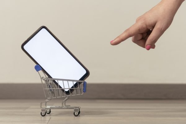 Na imagem com cor, dedo aponta para um carro de compras que tem um celular - Metrópoles