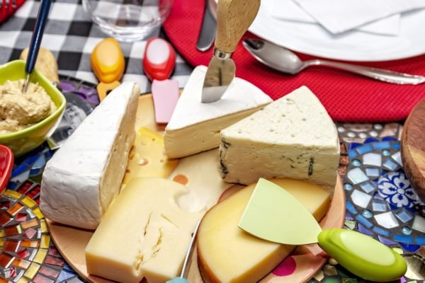 Sobre a mesa com toalha xadrez, vemos sobre uma tábua de vidro alguns queijos, como Gorgonzola, Brie, Provolone, Camembert e Estepe