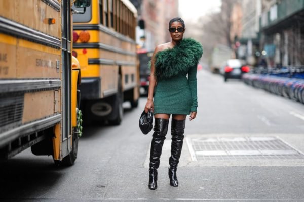 Em uma rua de Nova York, com ônibus amarelo passando ao lado, mulher negra posa. Ela usa botas pretas de cano alto e vestido verde assimétrico com plumas na parte de cima - Metrópoles