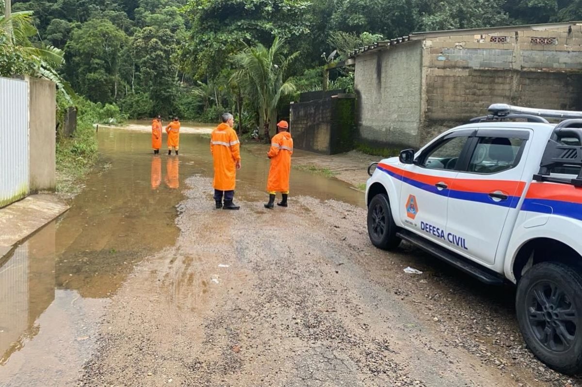 Três pessoas vestidas com uma capa laranja paradas na rua enquanto há um carro da Defesa Civil parado ao lado deles