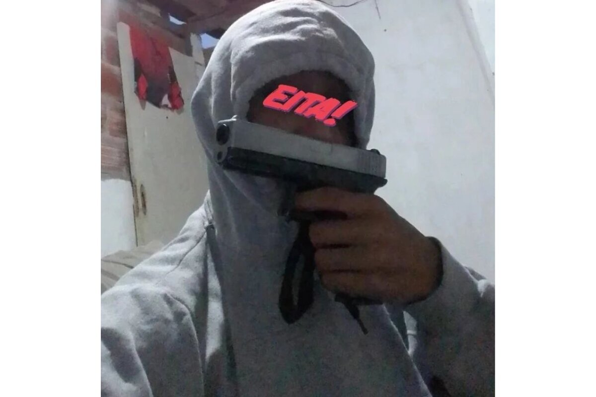 Foto colorida de jovem de moletom cinza claro, cobrindo o rosto com o capuz e uma pistola - Metrópoles