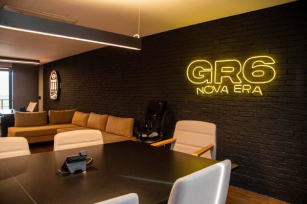 Sala de reuniões com mesa e sofás ao fundo, na parede preta de tijolinhos está uma logomarca "GR6 Nova Era" em neon amarelo