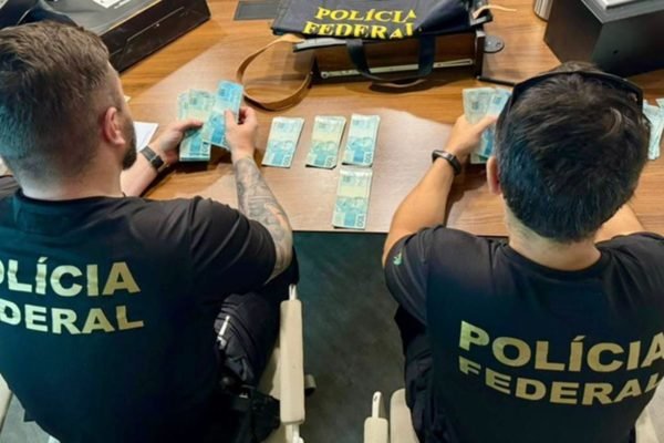 Imagem colorida das costas de dois policiais federais (PF) que estão conferindo cédulas de R$ 100 - Metrópoles