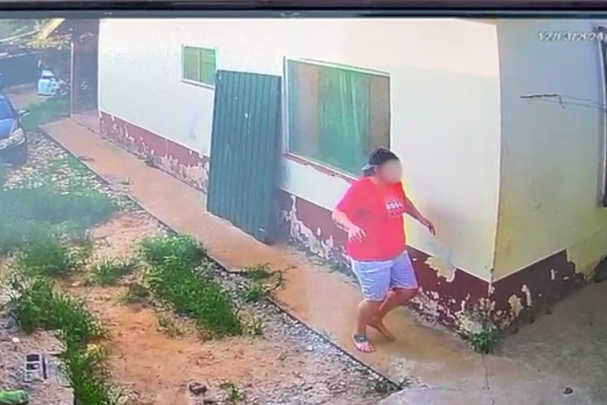 Homem com sobrepeso, usando camiseta vermelha, corre ao lado de casa em chão de terra batida - Metrópoles