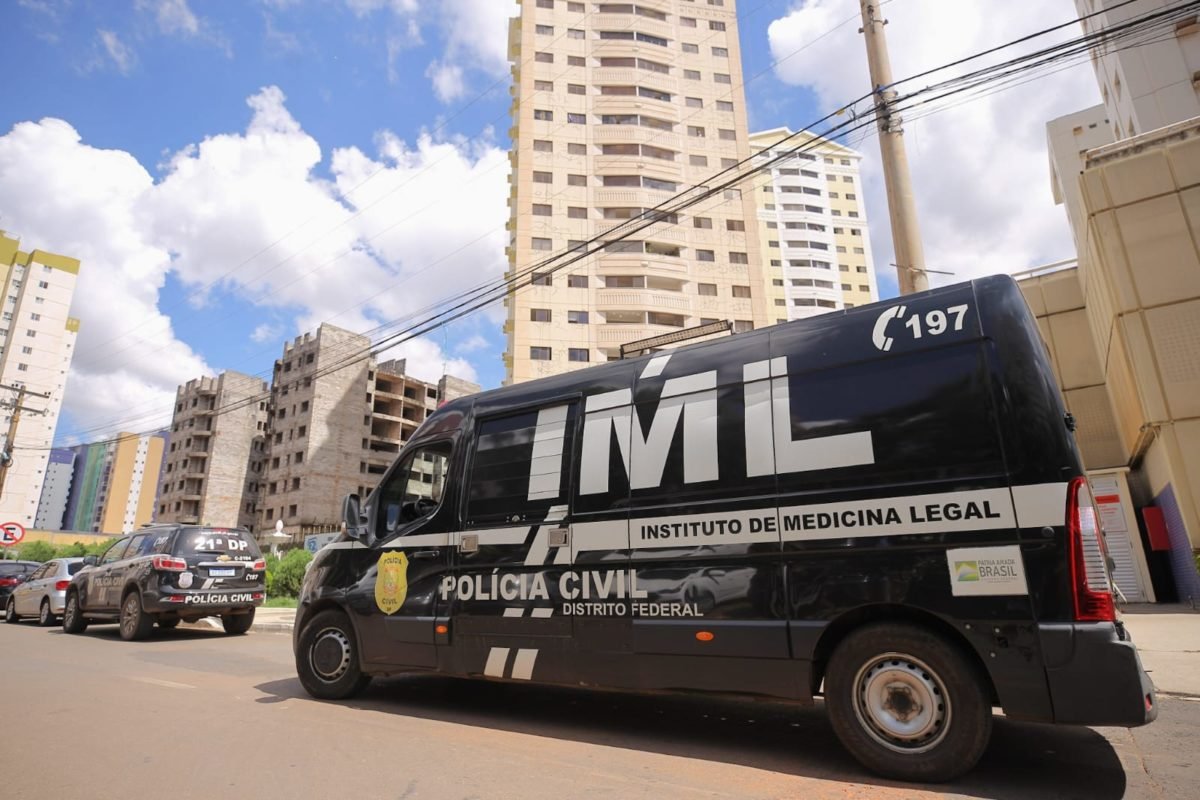 Foto colorida tirada de dia de uma van preta em frente à um prédio alto. Na lataria da van, estão as identificações do Instituto de Medicina Lega (IML).