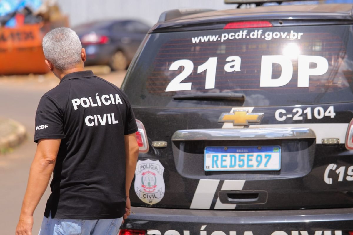 Foto colorida tirada de dia de homem de camiseta preta em frente à traseira de um carro preto. Tanto a camisa do homem, quanto o carro possuem a caracterização da Polícia Civil do Distrito Federal