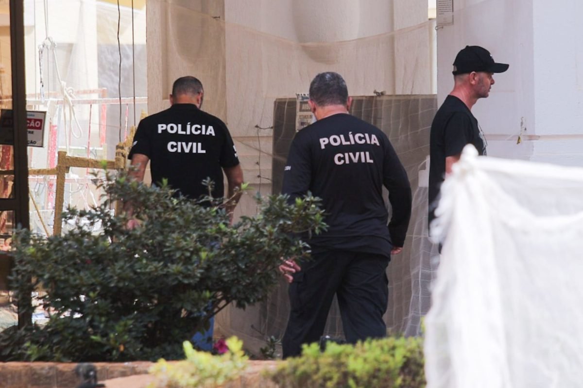 Foto colorida tirada de dia de três homens em meio a uma área interditada para obras. Dois dos homens apresentam uma camiseta com a identificação da Polícia Civil.
