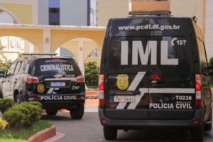 Foto colorida tirada de dia de um carro e uma van preta em frente à entrada de um prédio. Tanto o carro quanto a van apresentam as caracterizações da Polícia Civil do Distrito Federal.