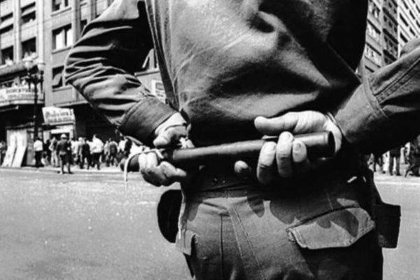 Imagem em preto e branco de policial fardado, de costas, segurando um cassetete. Ao fundo, manifestantes contra a ditadura militar