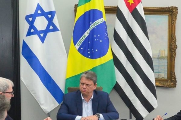 Tarcísio de Freitas sentado em uma mesa e ao fundo três bandeiras: uma de Israel, uma do Brasil e outra do Estado de SP - Metrópoles