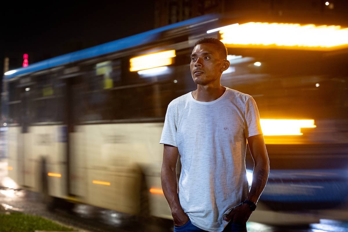 Foto de homem pardo e blusa branca com ônibus passando ao fundo