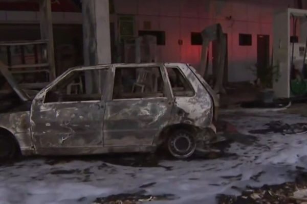 Imagem colorida de Fiat Uno branco destruído por incêndio após o carro bater em bomba de posto de combustível. Ao redor, no chão, h[a espuma branca usada para conter o fogo.