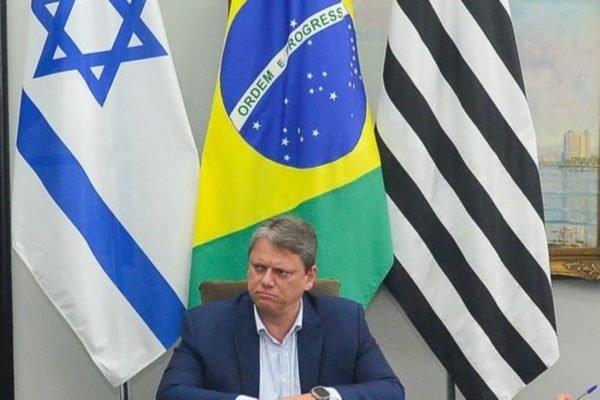 Foto colorida do governador Tarcísio de Freitas vestindo terno azul e camisa clara com bandeiras de Israel, Brasil e São Paulo ao fundo - Metrópoles