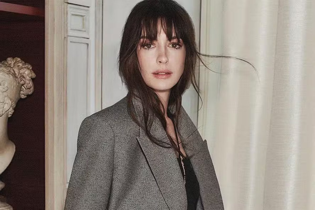 Anne Hathaway em foto colorida. Ela é uma mulher branca, de cabelo castanho. Usa uma blusa preta e um blazer cinza