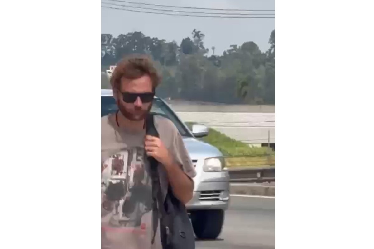 Homem de barba e óculos escuros caminha na lateral de rodovia segurando mochila somente no ombro esquerdo - Metrópoles