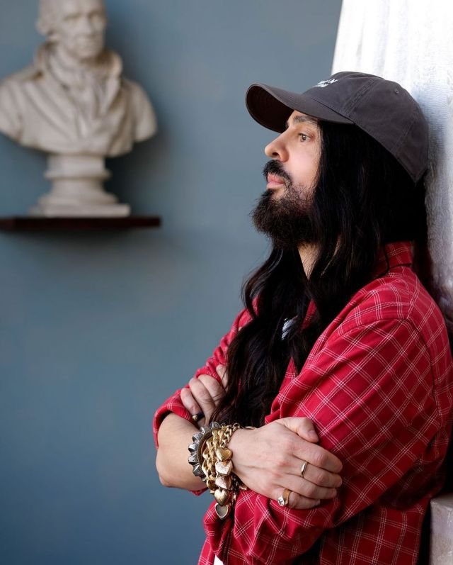 Alessandro Michele, homem branco com barba e cabelo longo, usando boné. Ele está de braços cruzados e encostado na parede - Metrópoles